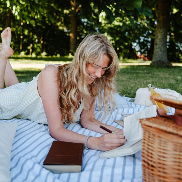Person in sundress lying on picnic blanket writing in journal.